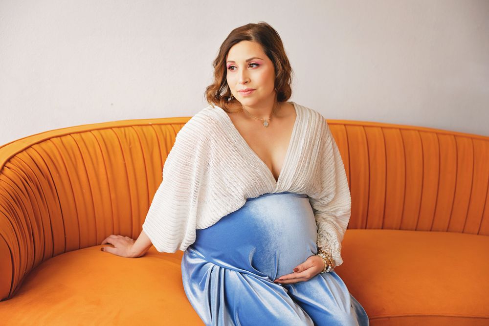 Maternity pose on orange velvet couch wearing cream sweater and blue dress.