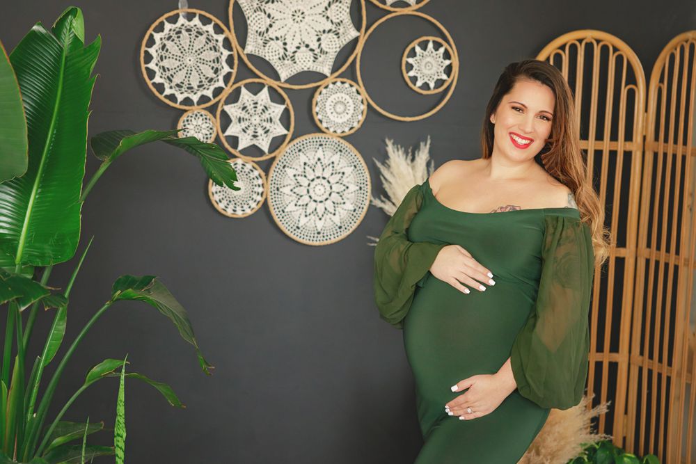 Maternity portrait in green dress with bohemian decor and plants against dark wall.