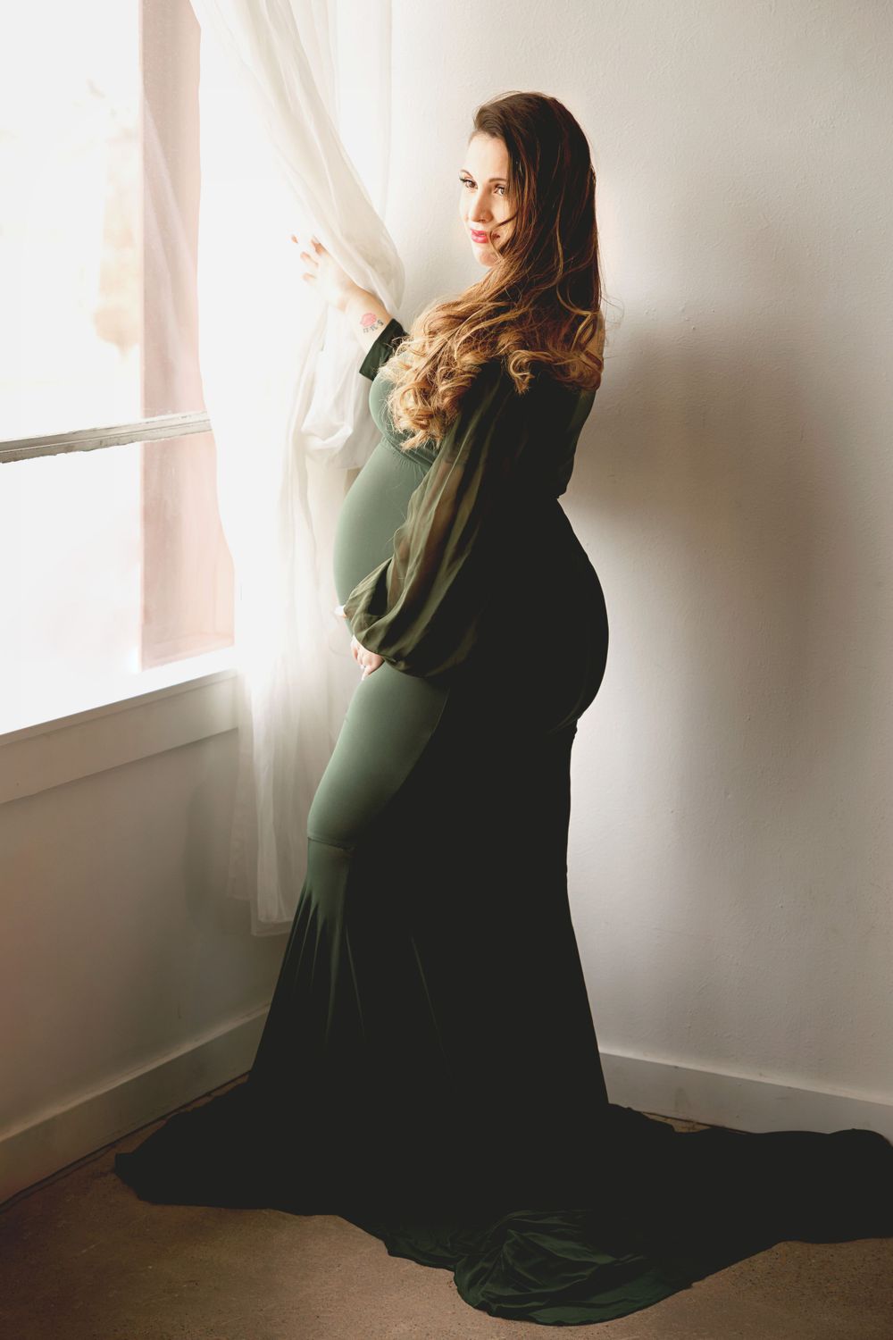 Maternity silhouette in flowing dark gown standing by window in soft natural light.