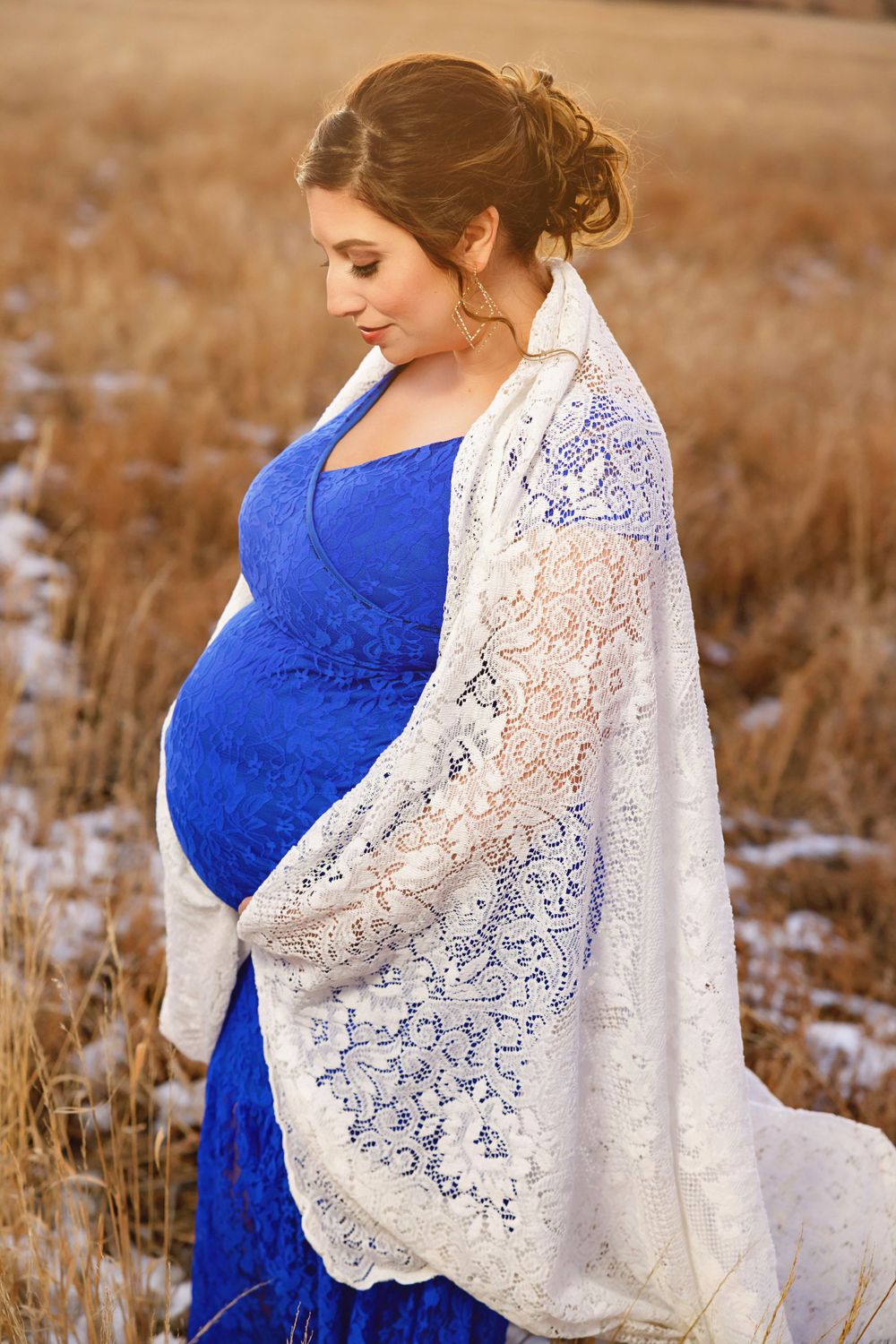 Expectant mother in royal blue dress with white lace shawl poses in snowy winter field.
