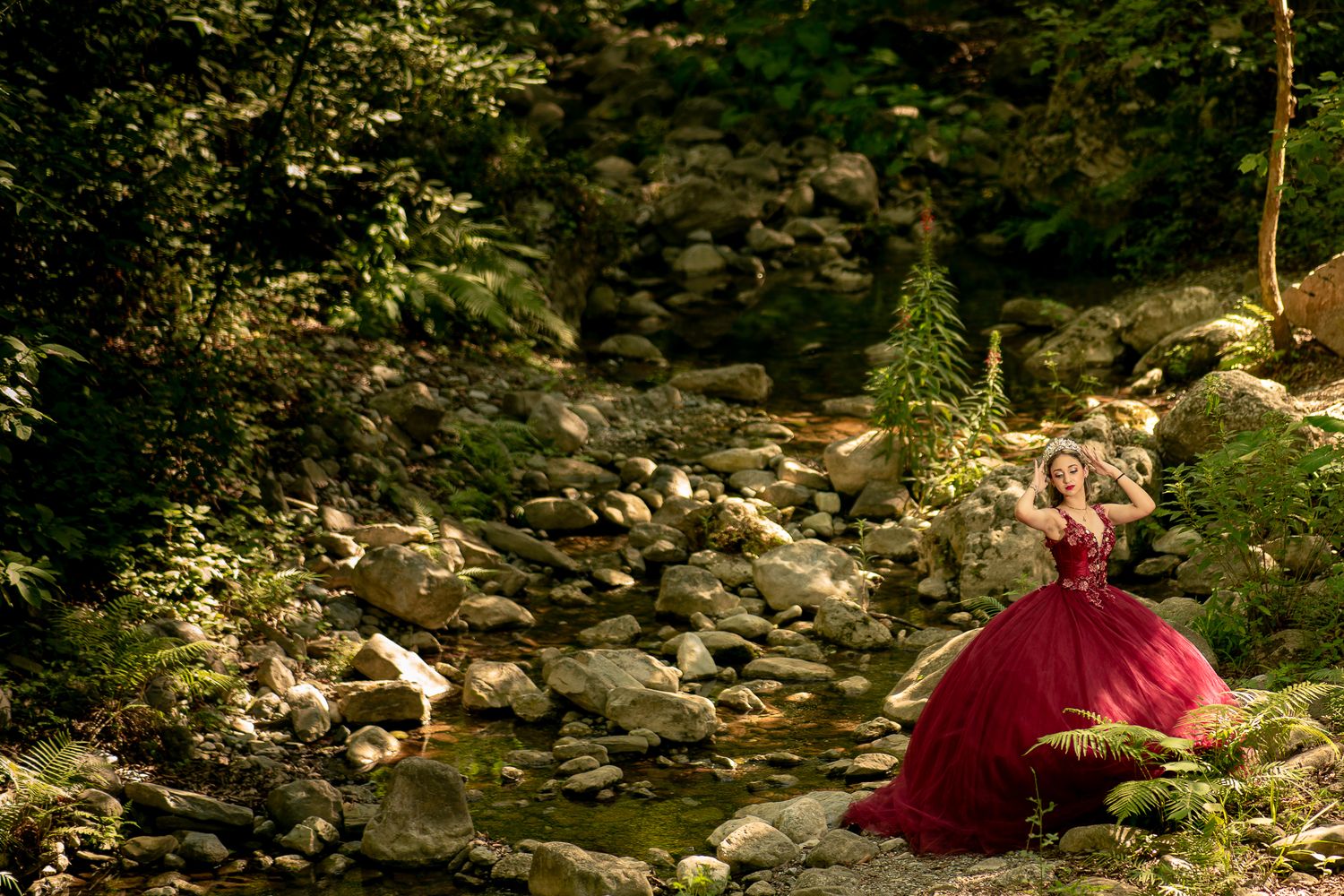 Person in burgundy outfit sitting by rocky stream in forest setting.