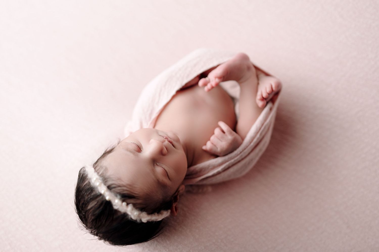 Newborn baby sleeping peacefully on white fabric with soft pink background.