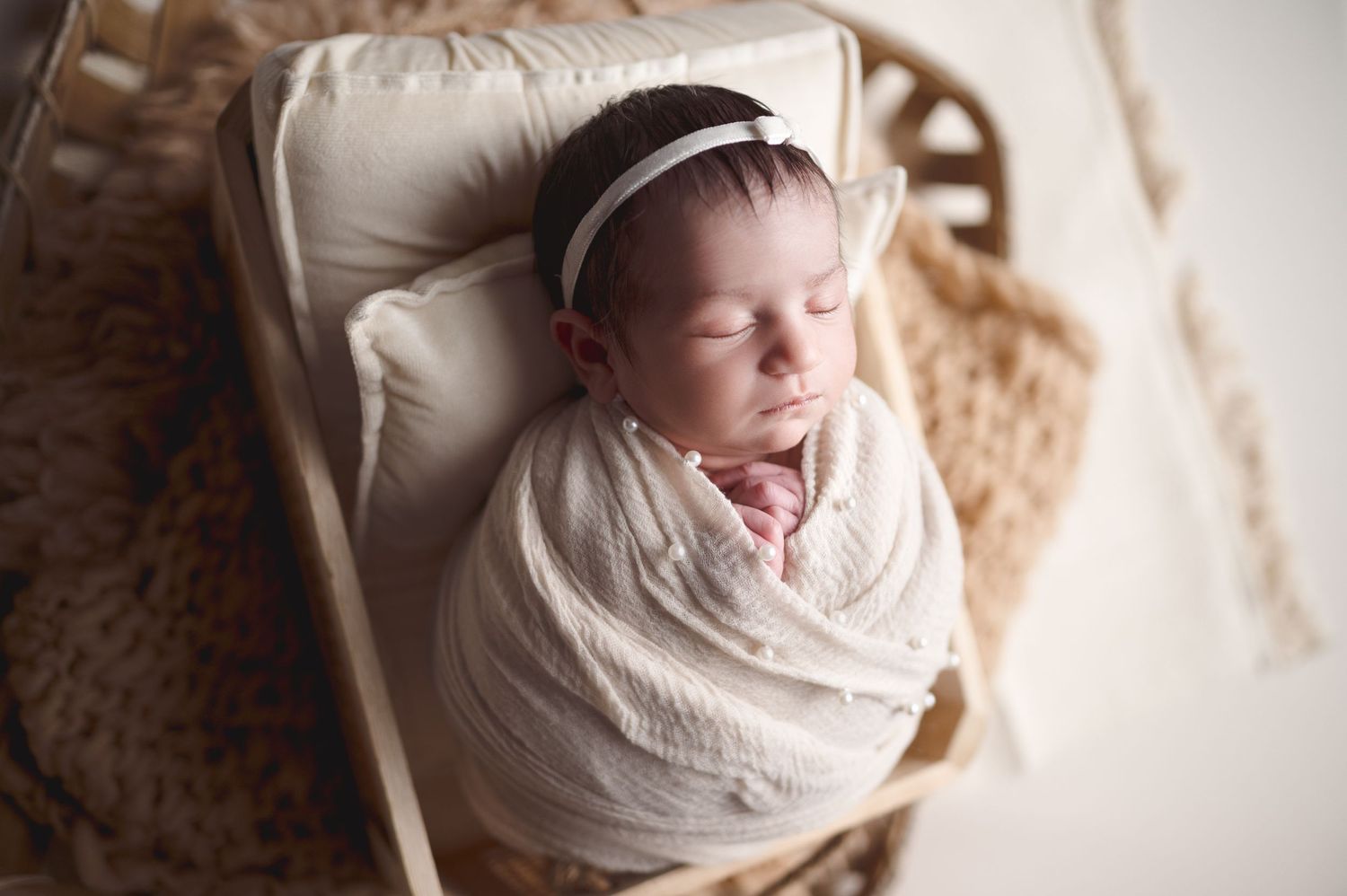 Peaceful newborn baby sleeps while wrapped in white swaddle on textured cream background.