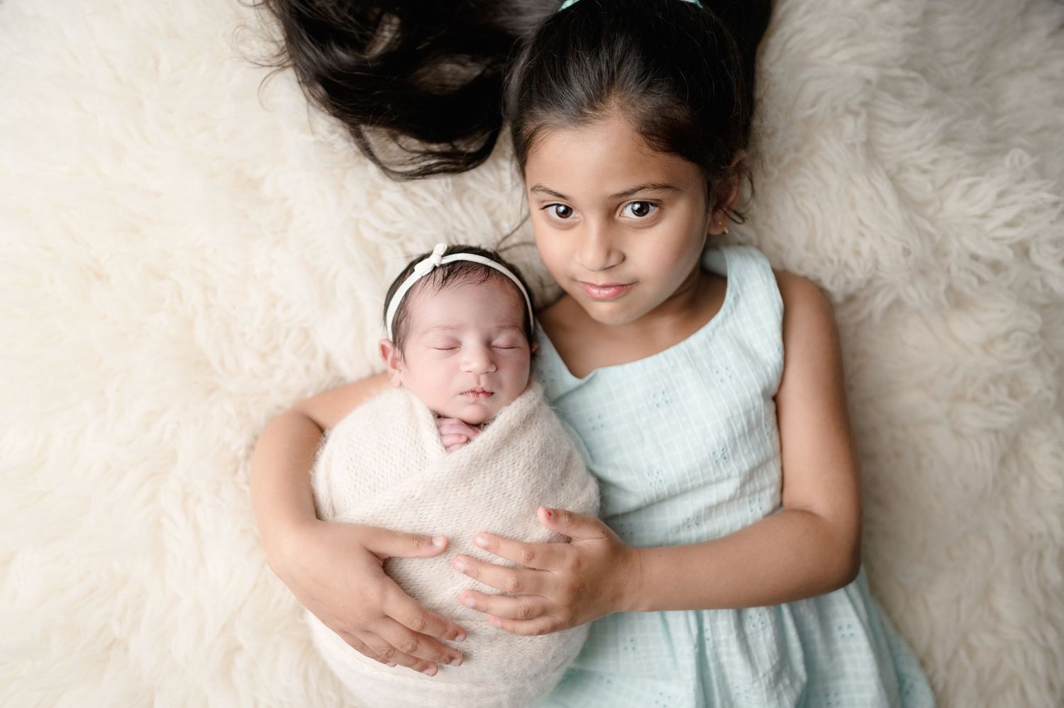 Siblings bonding moment as older sister lies next to swaddled newborn on white fur backdrop.