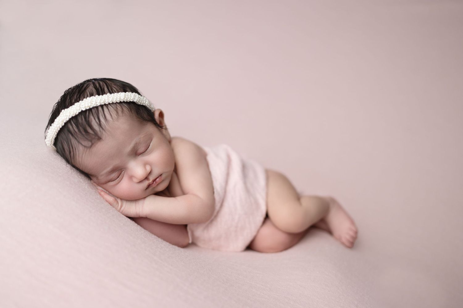 Artistic newborn portrait with white headband against soft pink background.