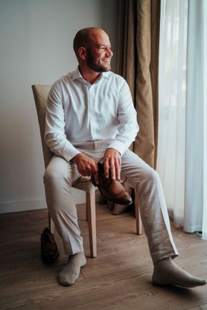 A person in all white clothing sits relaxed on a wooden chair by a window.