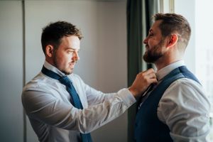 Two people in formal attire helping with tie adjustments near a window.