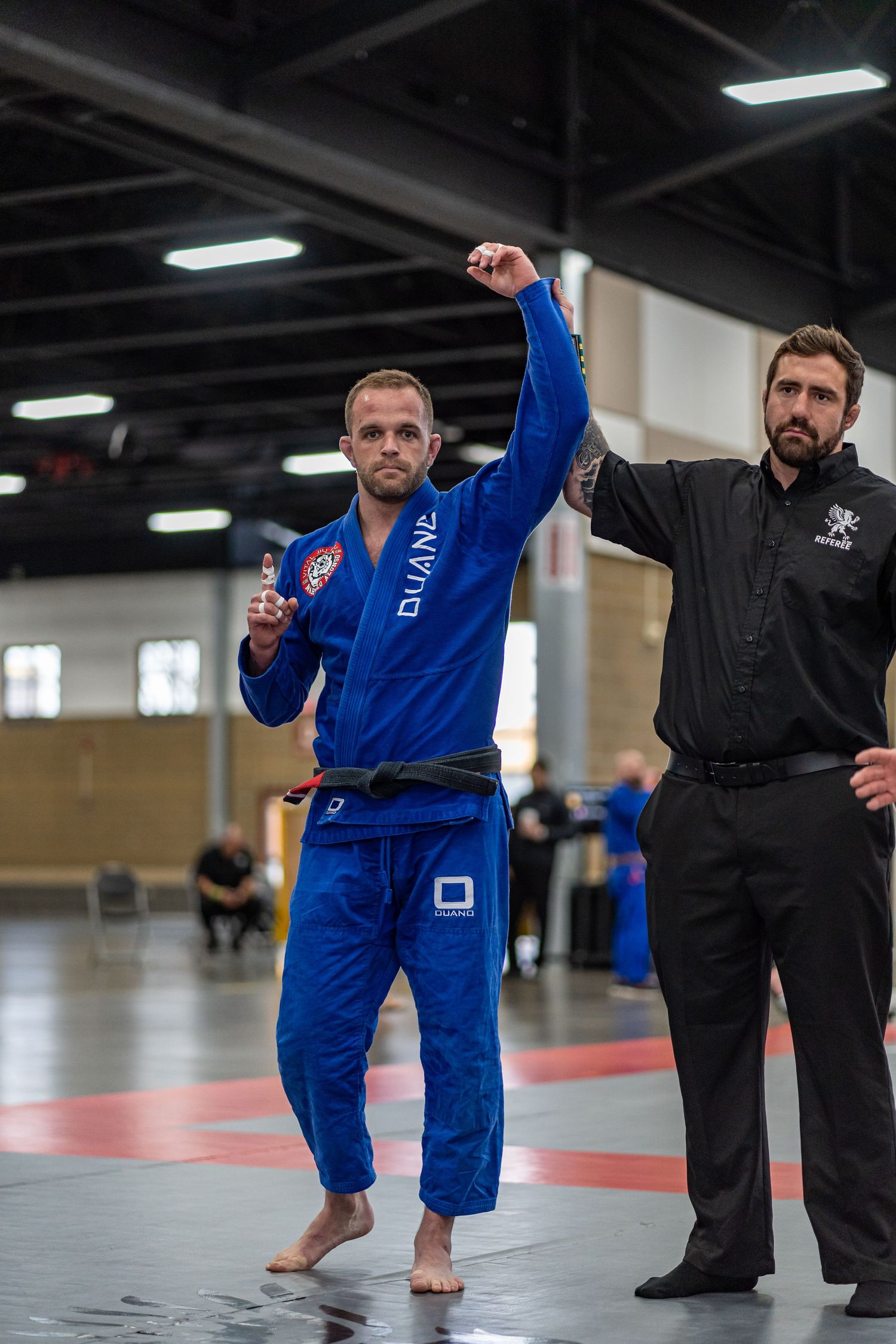 A competitor in a blue gi celebrates victory with raised arms as a referee signals the win.