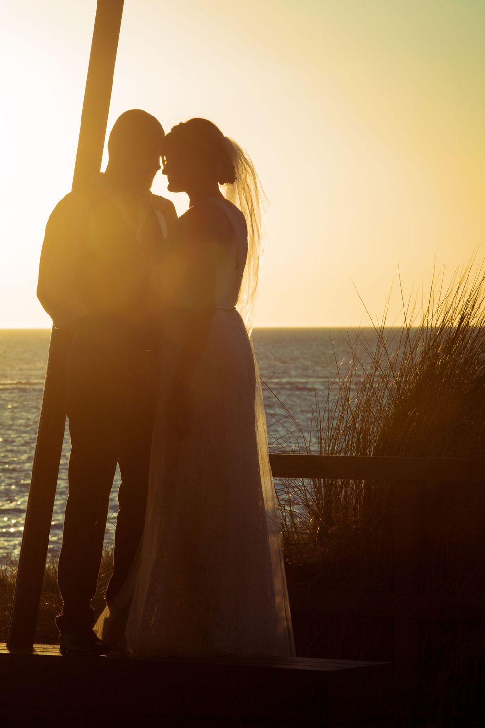 Silhouetted couple stands together against golden sunset sky over ocean.