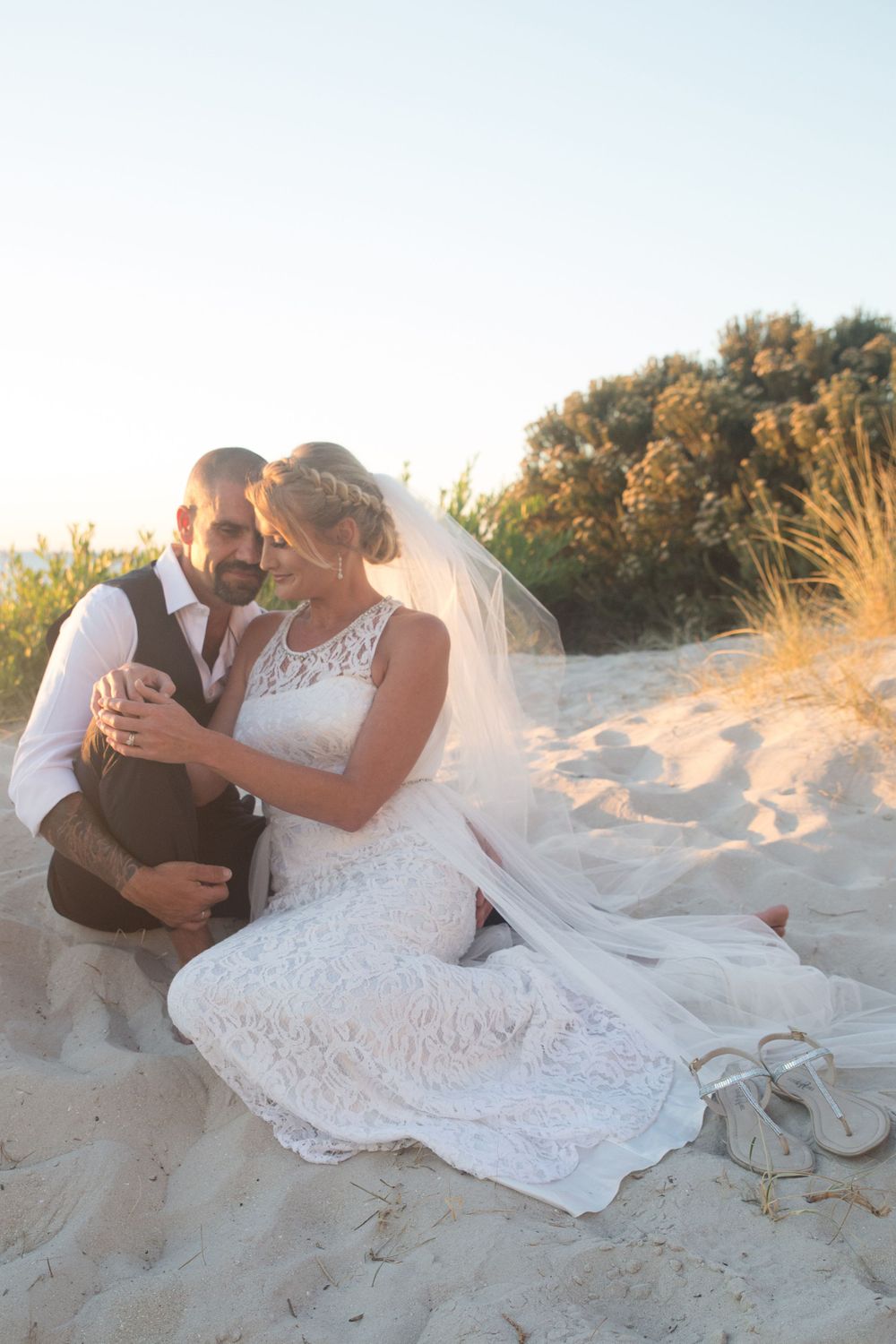 Intimate beach wedding photoshoot with couple lounging in white attire on sandy dunes at sunset.