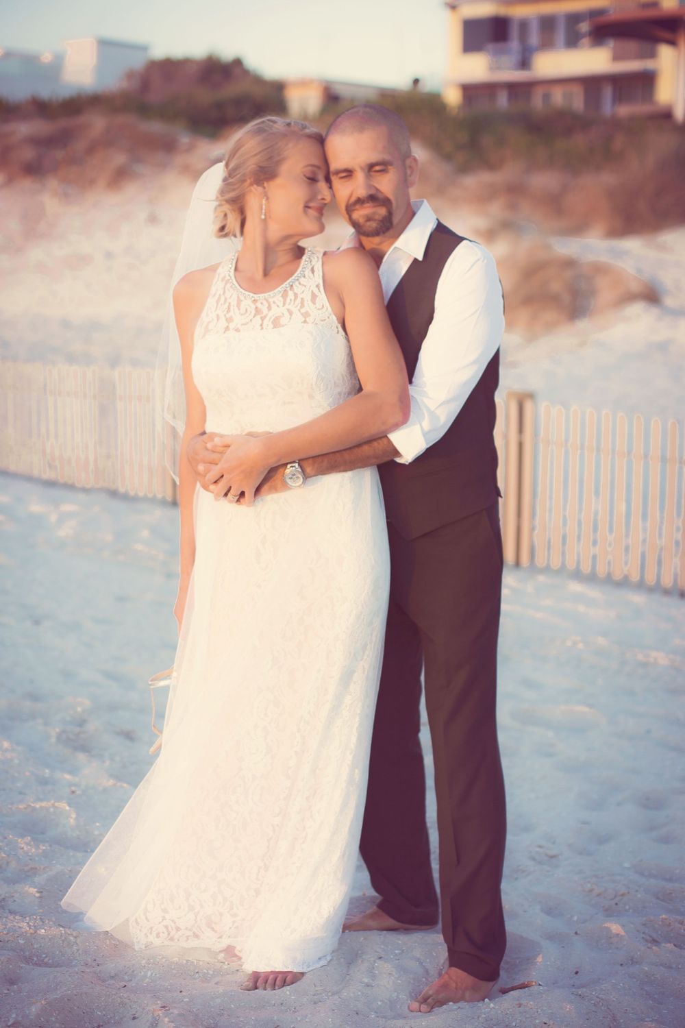 Beachside wedding photos show couple embracing in soft focus with white picket fence backdrop.