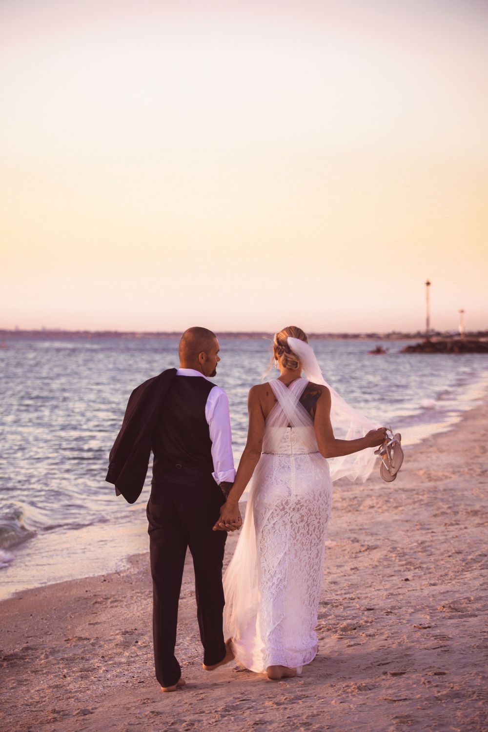 Newlyweds walking hand in hand on beach at sunset, bride in lace gown and groom in dark suit.