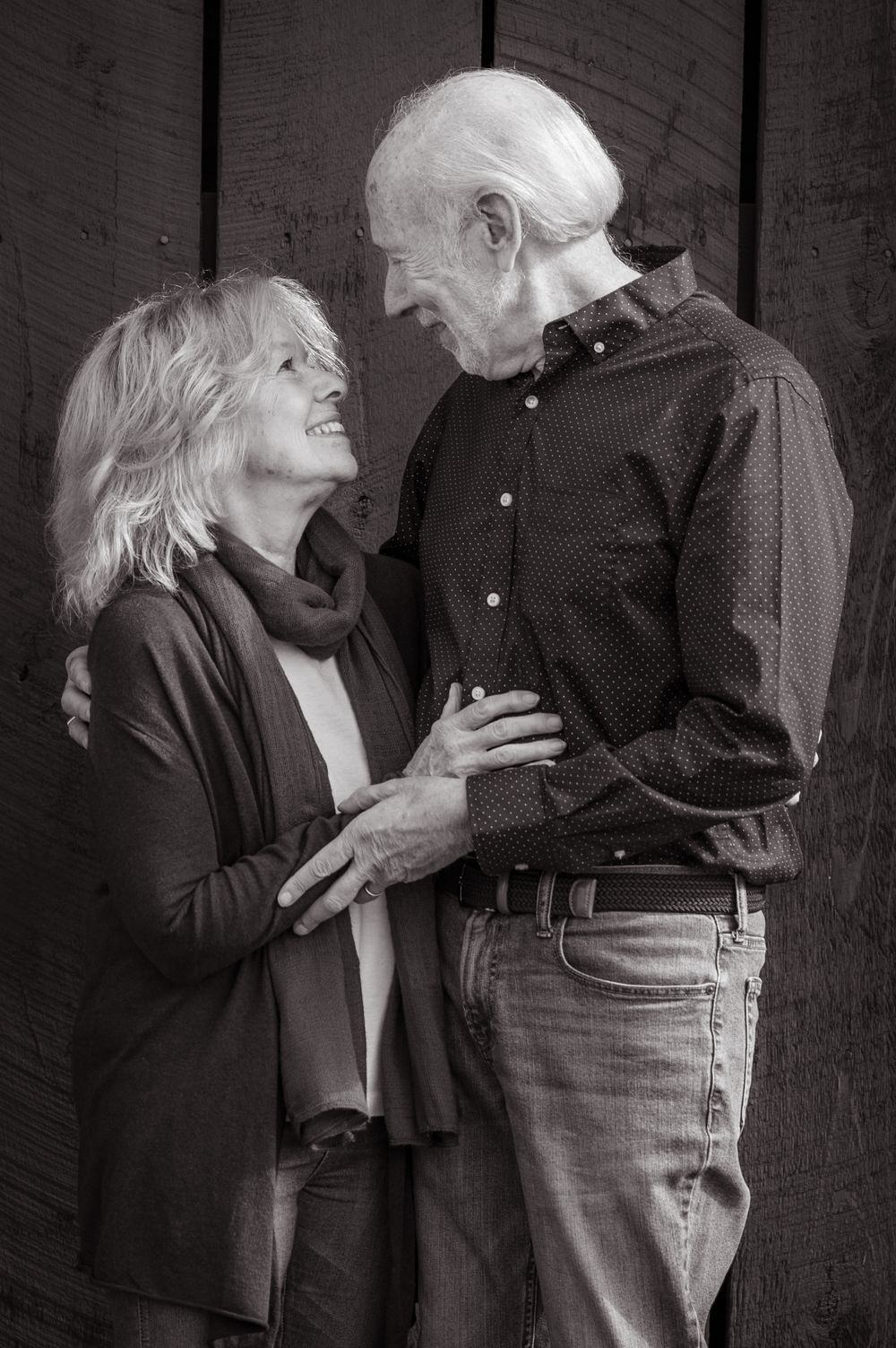 An elderly couple share a tender moment against a rustic wooden wall in black and white.