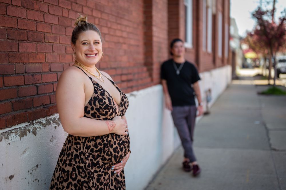 An expectant mother in a leopard print dress poses near a brick wall while her partner admires in the background.