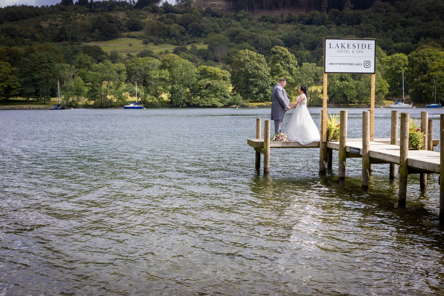 Bride sitting on lakeside dock with mountains and trees in background.