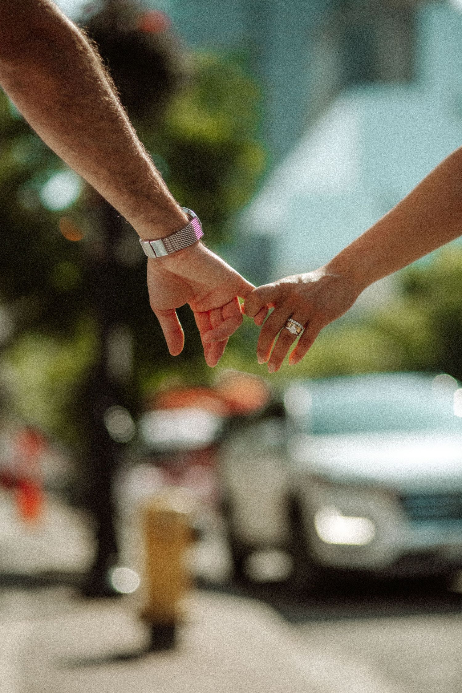 Close-up shot of two hands holding each other wearing engagement rings on a sunny street.