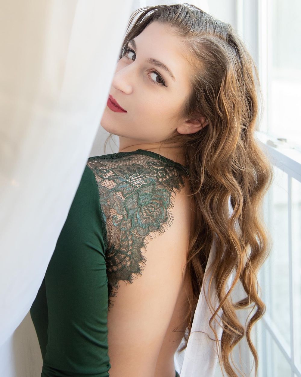 Person in dark green dress with lace detail poses by window.