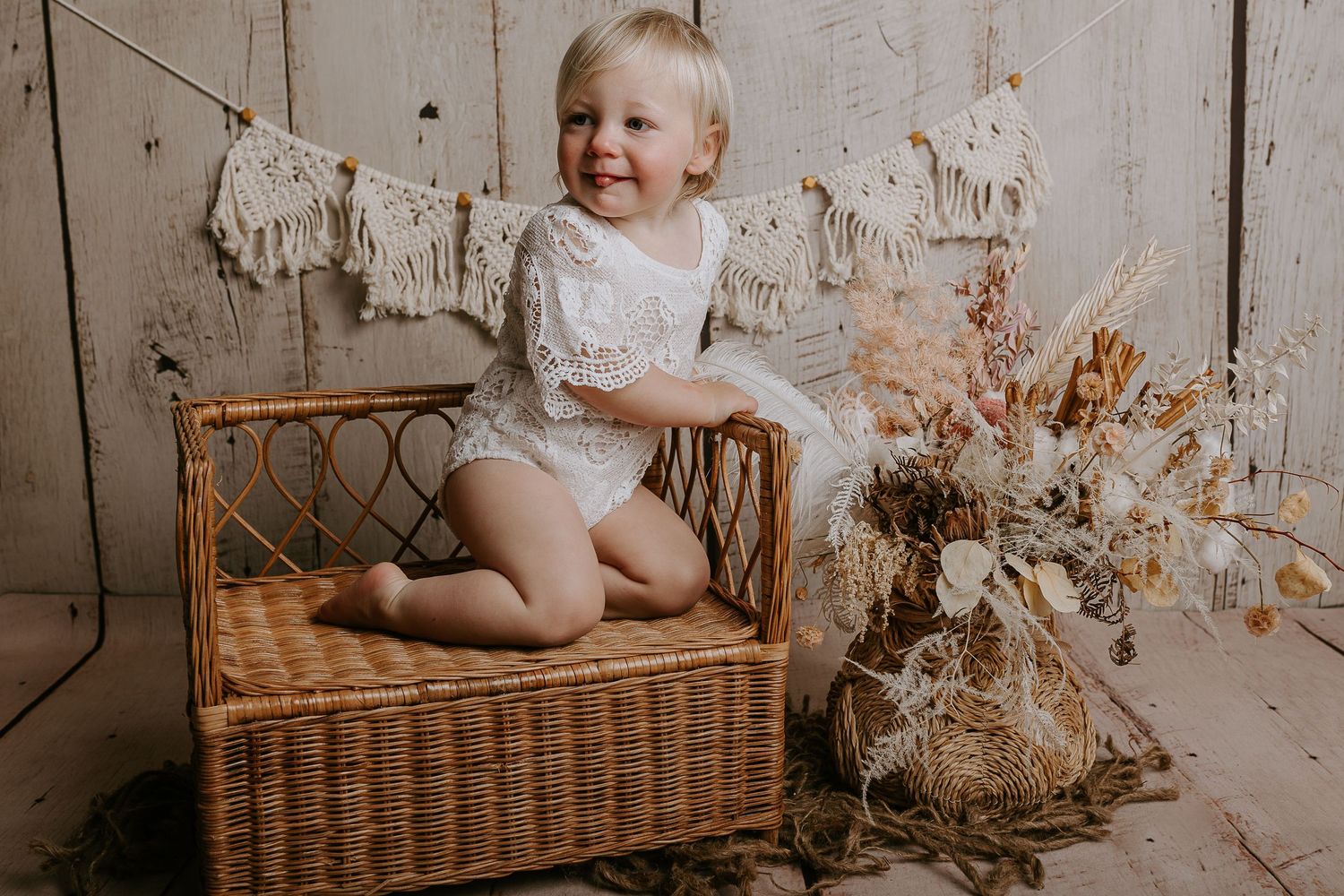 Smiling baby sits in wicker basket surrounded by dried flowers against rustic white wooden backdrop.