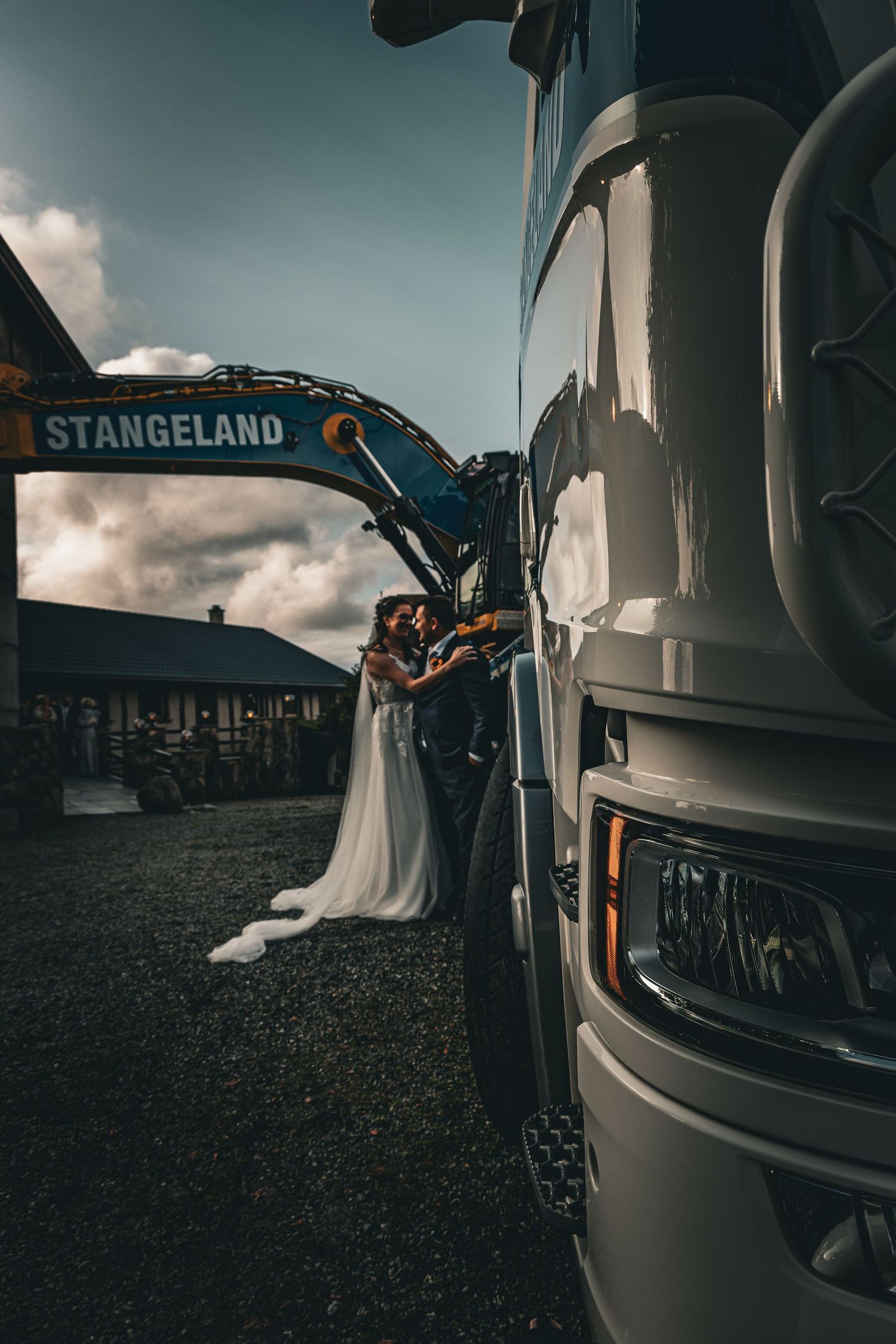 Bride and groom sharing kiss beside large truck with excavator at wedding venue.