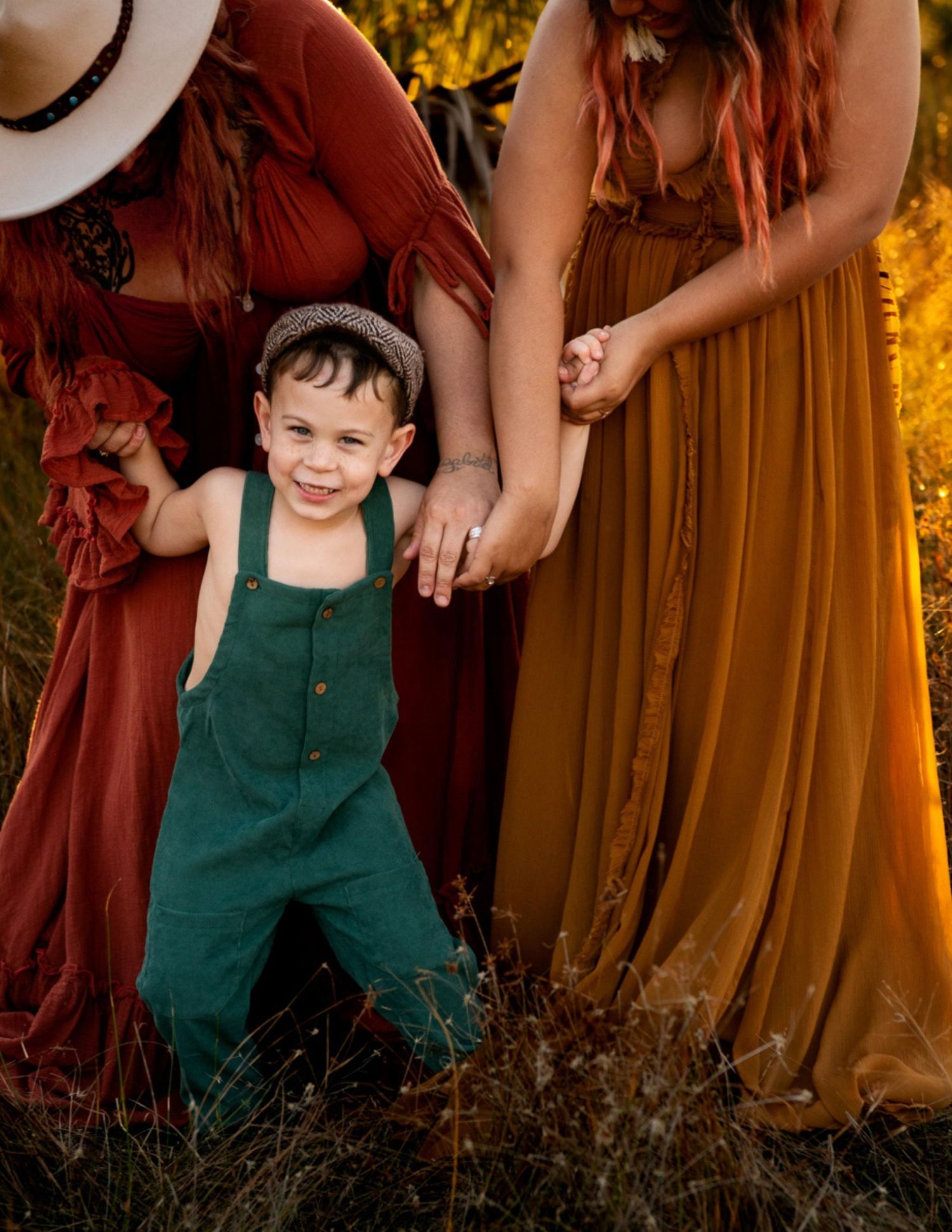 Smiling toddler boy in green overalls holds hands with two women in rust and mustard boho dresses outdoors.