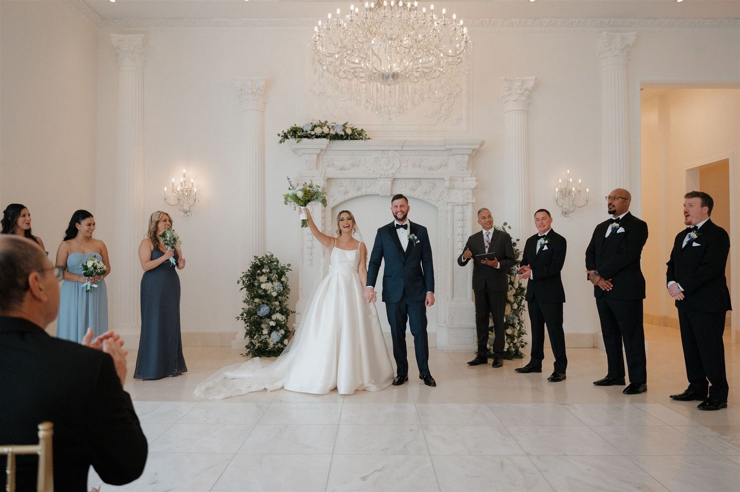 Wedding party stands in formation during ceremony in an elegant white room with crystal chandelier.
