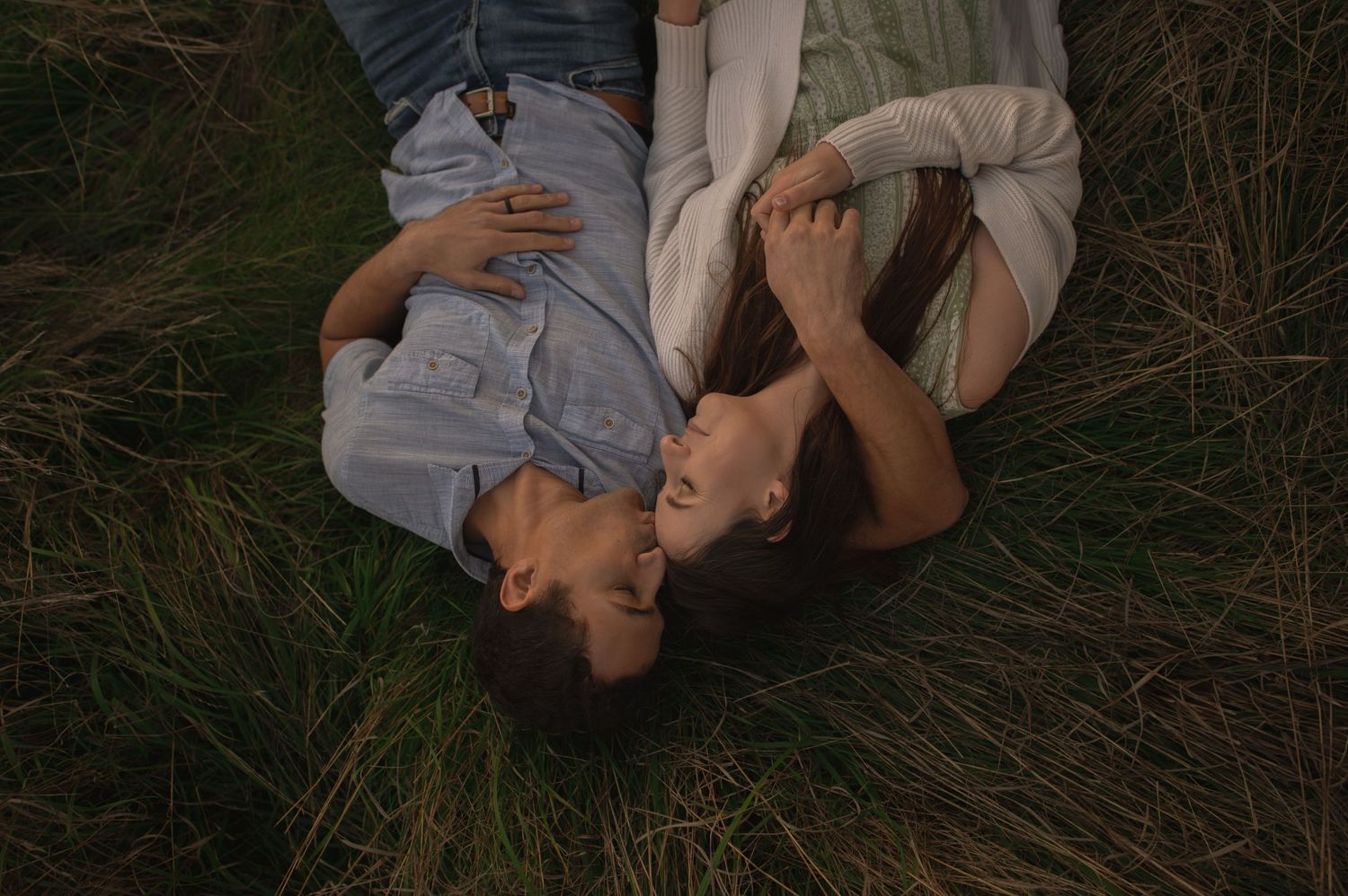 Two people laying in grass together at sunset in a romantic pose.