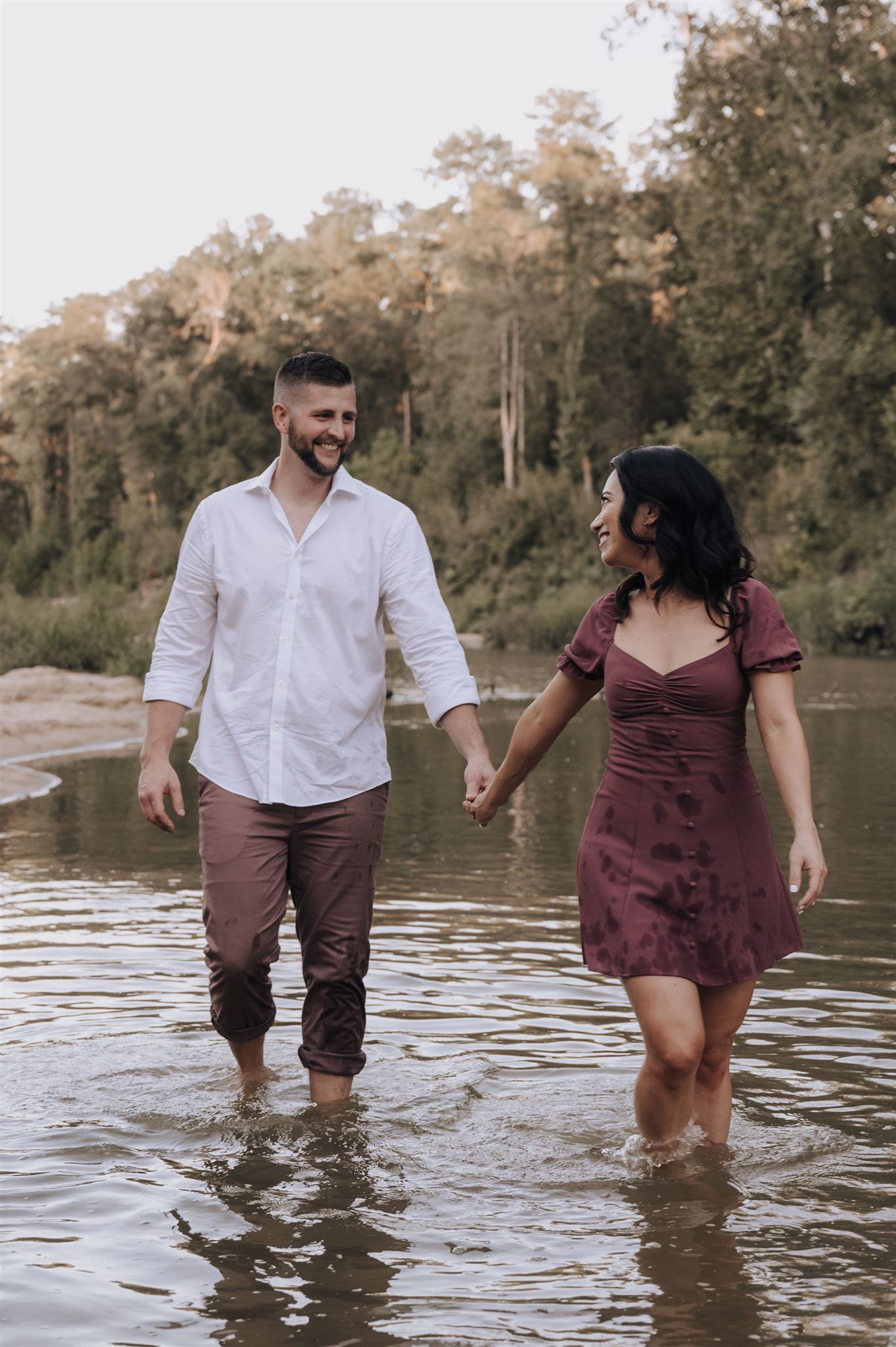 Couple holding hands while walking through shallow river water in coordinated burgundy and white outfits.