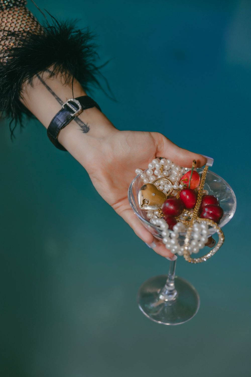 Hand holding martini glass decorated with pearls and cherries against teal background.