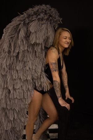 Model wearing black lingerie poses with large gray feathered wings in studio setting.
