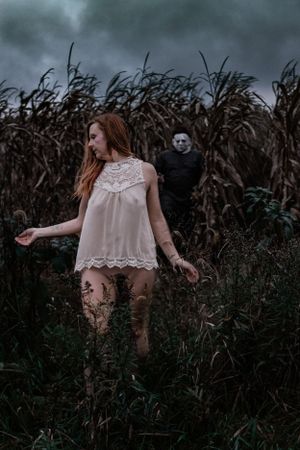 Eerie scene with white dress in cornfield stalked by masked figure.