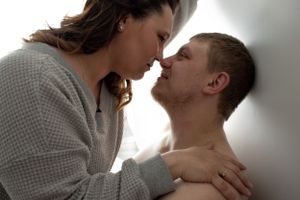 Intimate couple in soft lighting share romantic moment against white wall.