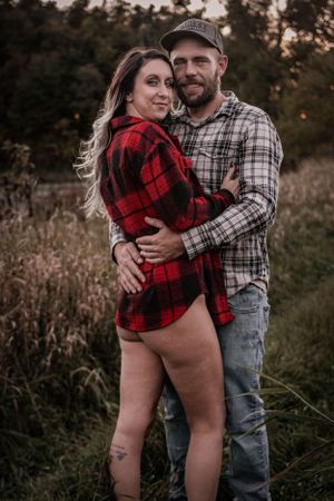 Intimate couple wearing matching plaid shirts embrace in tall grass at dusk.