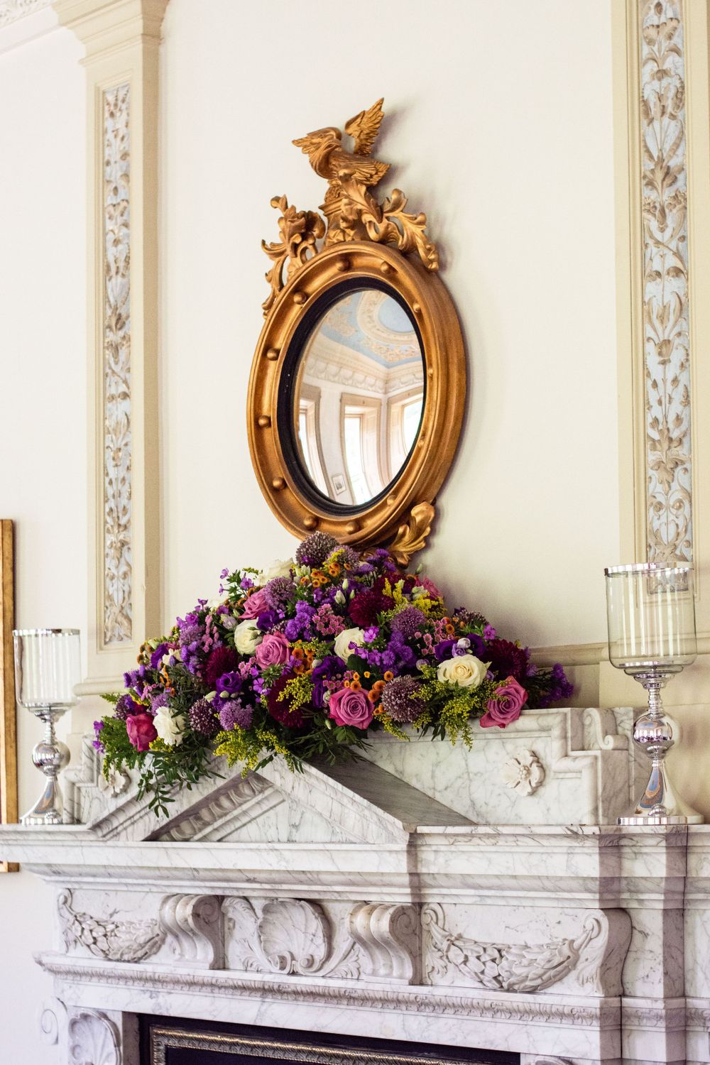 Ornate gold mirror above fireplace with vibrant purple and pink floral arrangement.