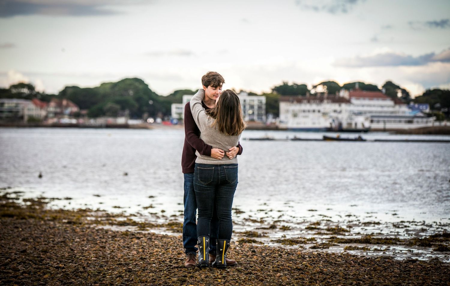 Couple sitting by the waterfront with boats and houses visible in the distance.