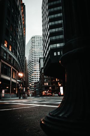 Low angle view of urban city street canyon with tall skyscrapers and glowing lights at twilight.