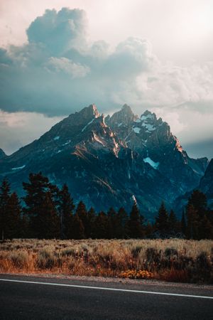 Scenic view of jagged mountain peaks with snow, pine trees, and a road in the foreground.