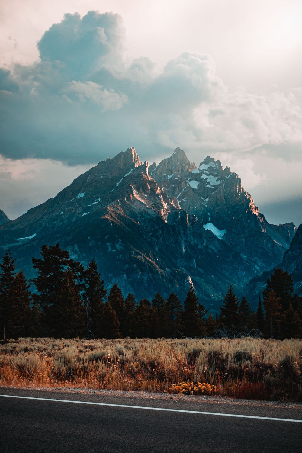 Scenic view of jagged mountain peaks with snow, pine trees, and a road in the foreground.
