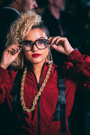Stylish woman in red jacket adjusting oversized glasses with gold chain necklace.