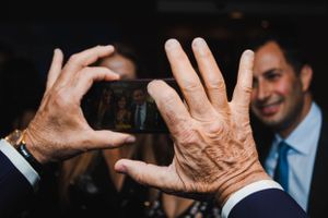 Elderly hands holding smartphone at event with blurred guests in background.