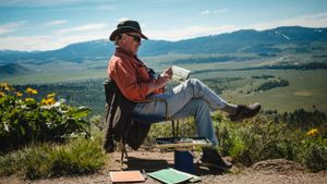 Man sitting in chair sketching scenic mountain valley landscape on hilltop.