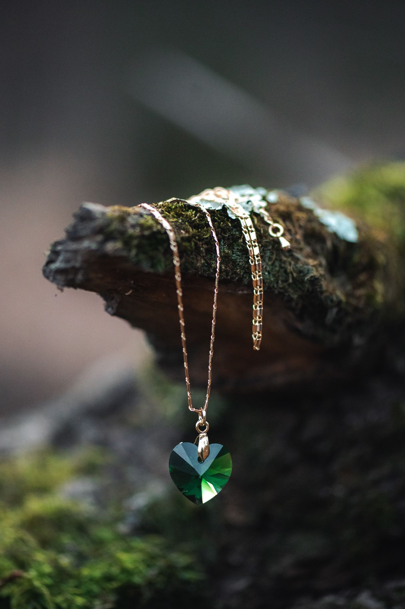 A delicate green crystal heart pendant on a gold chain rests on a mossy log in nature.