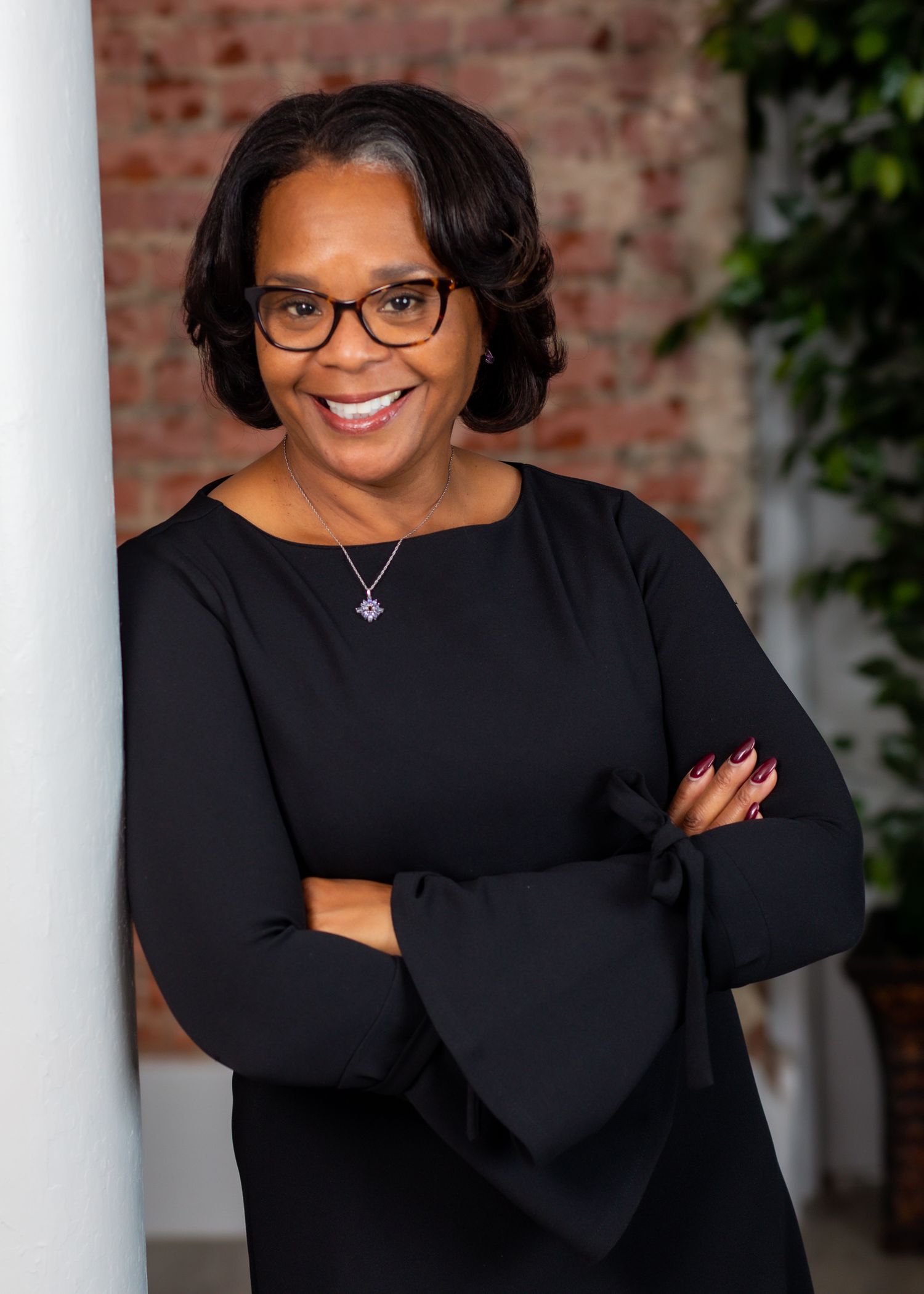 Professional portrait of person in black bell-sleeve dress against brick wall background.