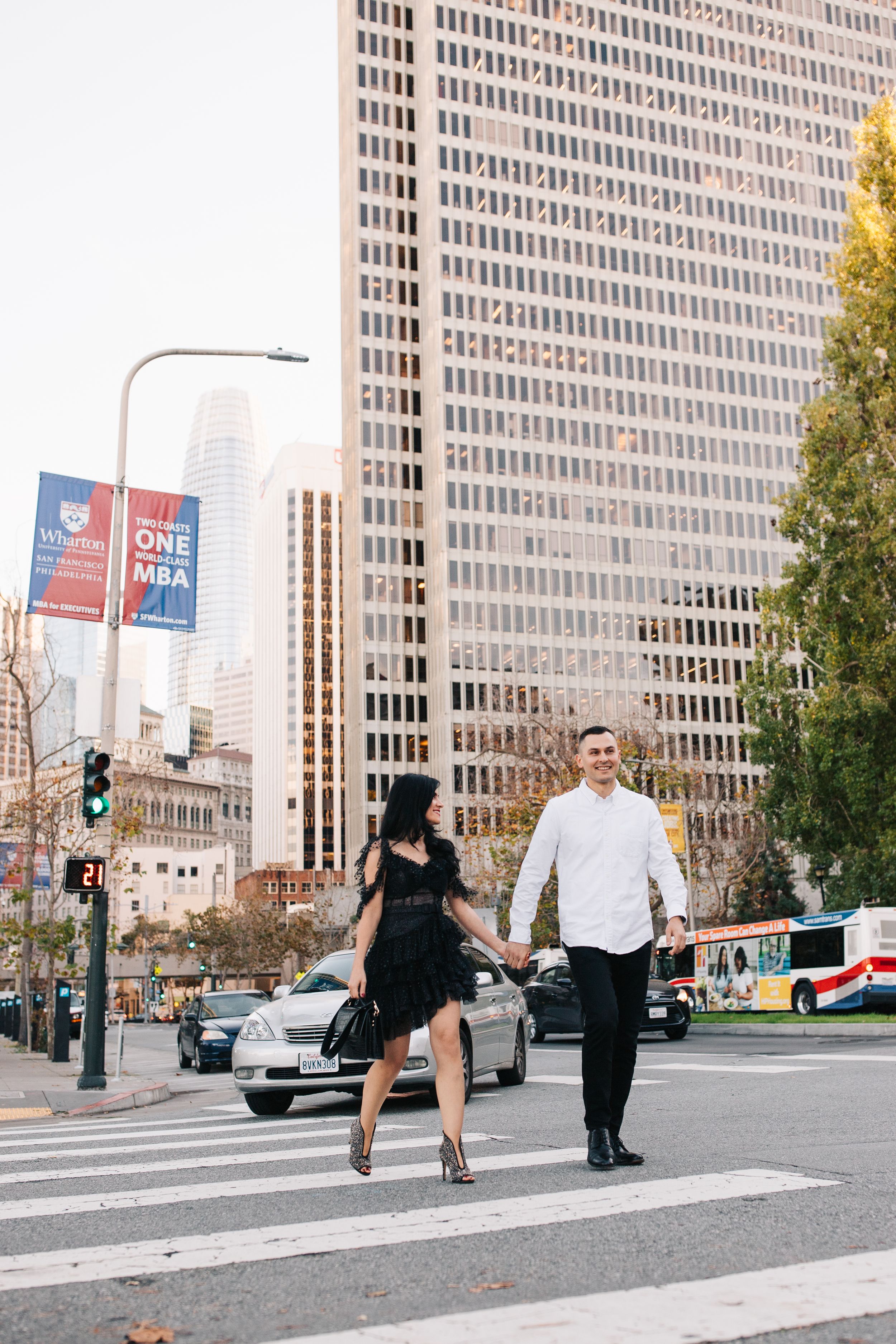 A couple walking across an urban crosswalk in downtown San Francisco with tall buildings behind them.