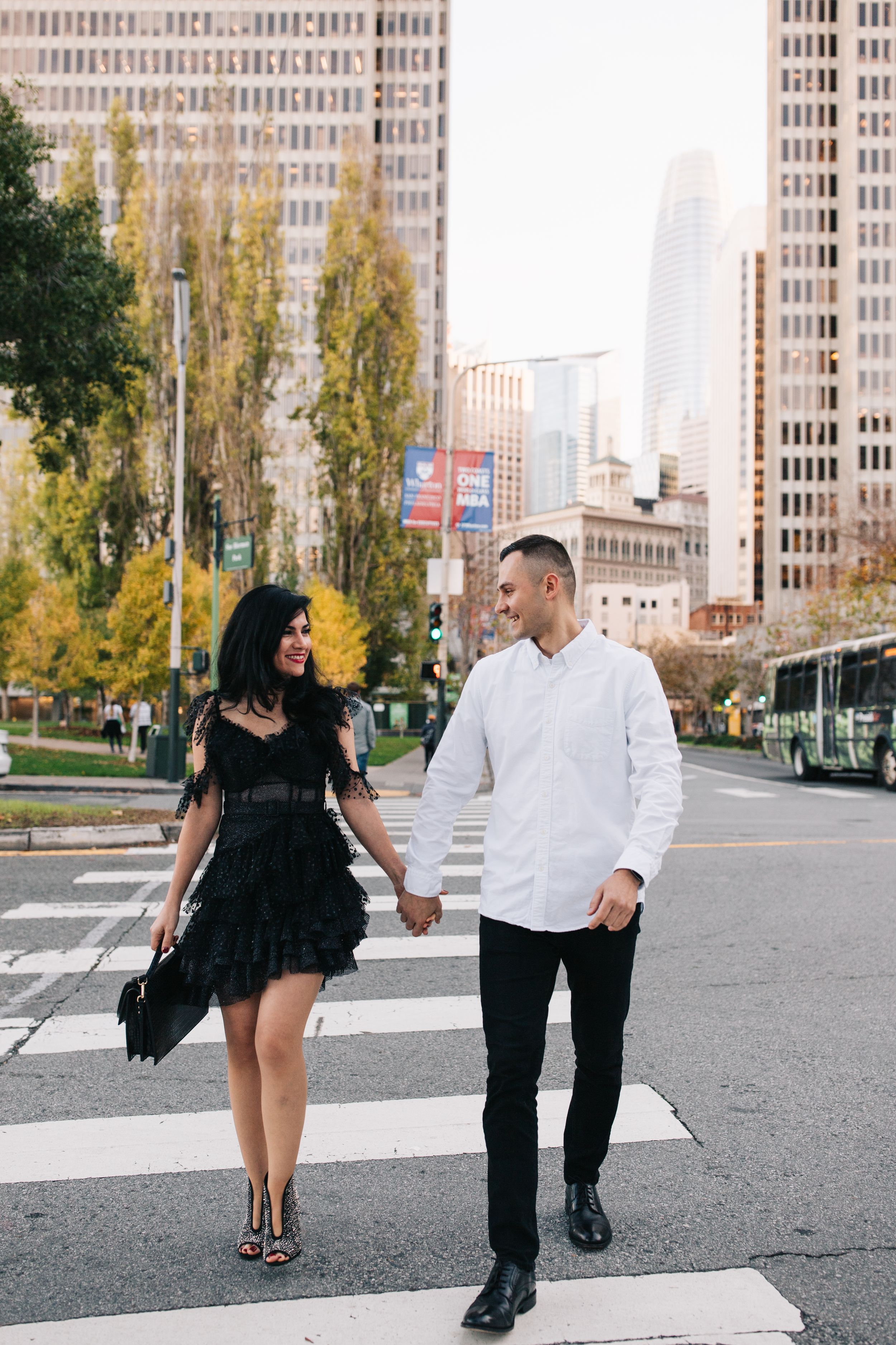 A stylishly dressed pair holding hands while crossing a city street with autumn trees and skyscrapers.