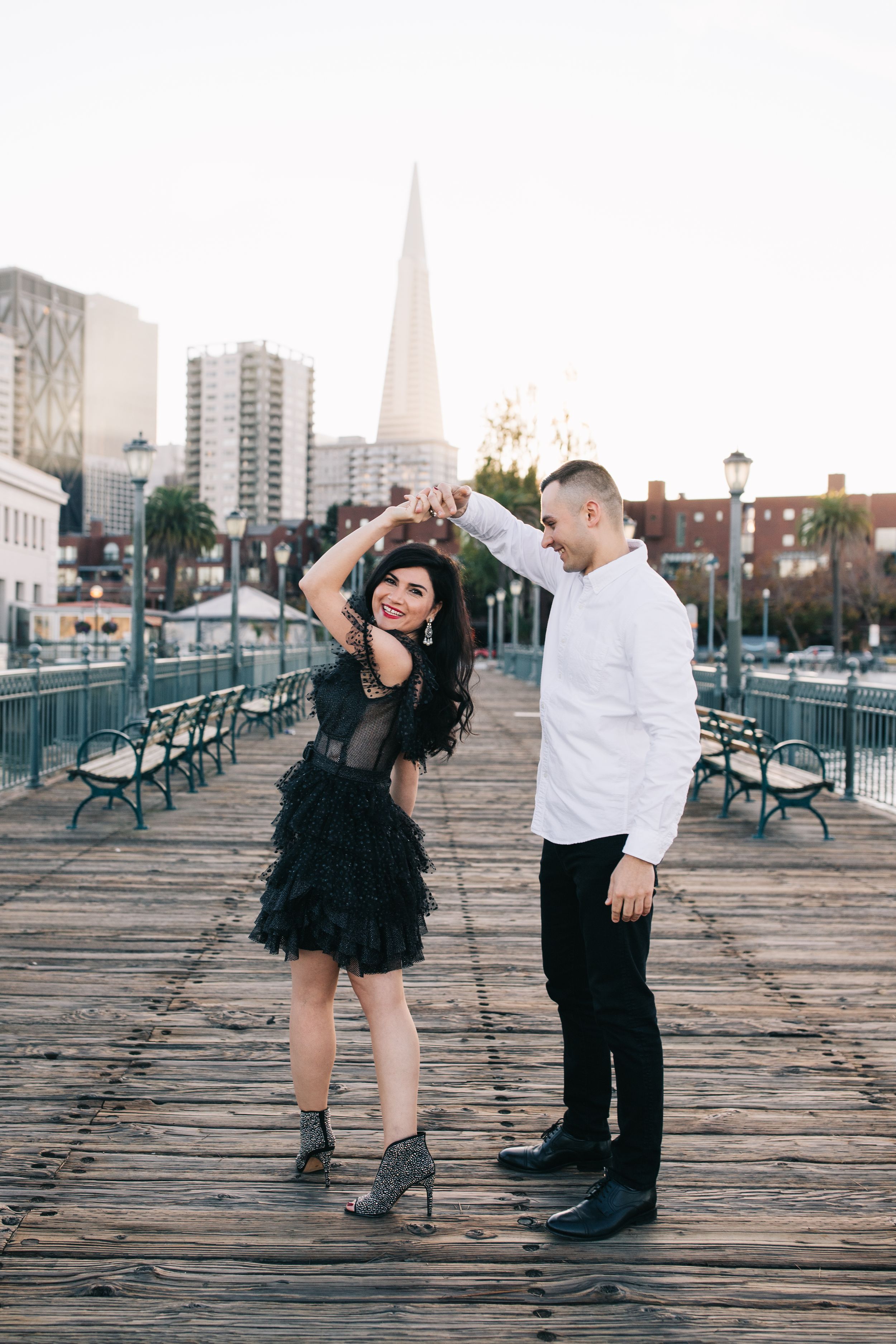 A couple in formal attire poses playfully on a wooden pier with city buildings in the distance.