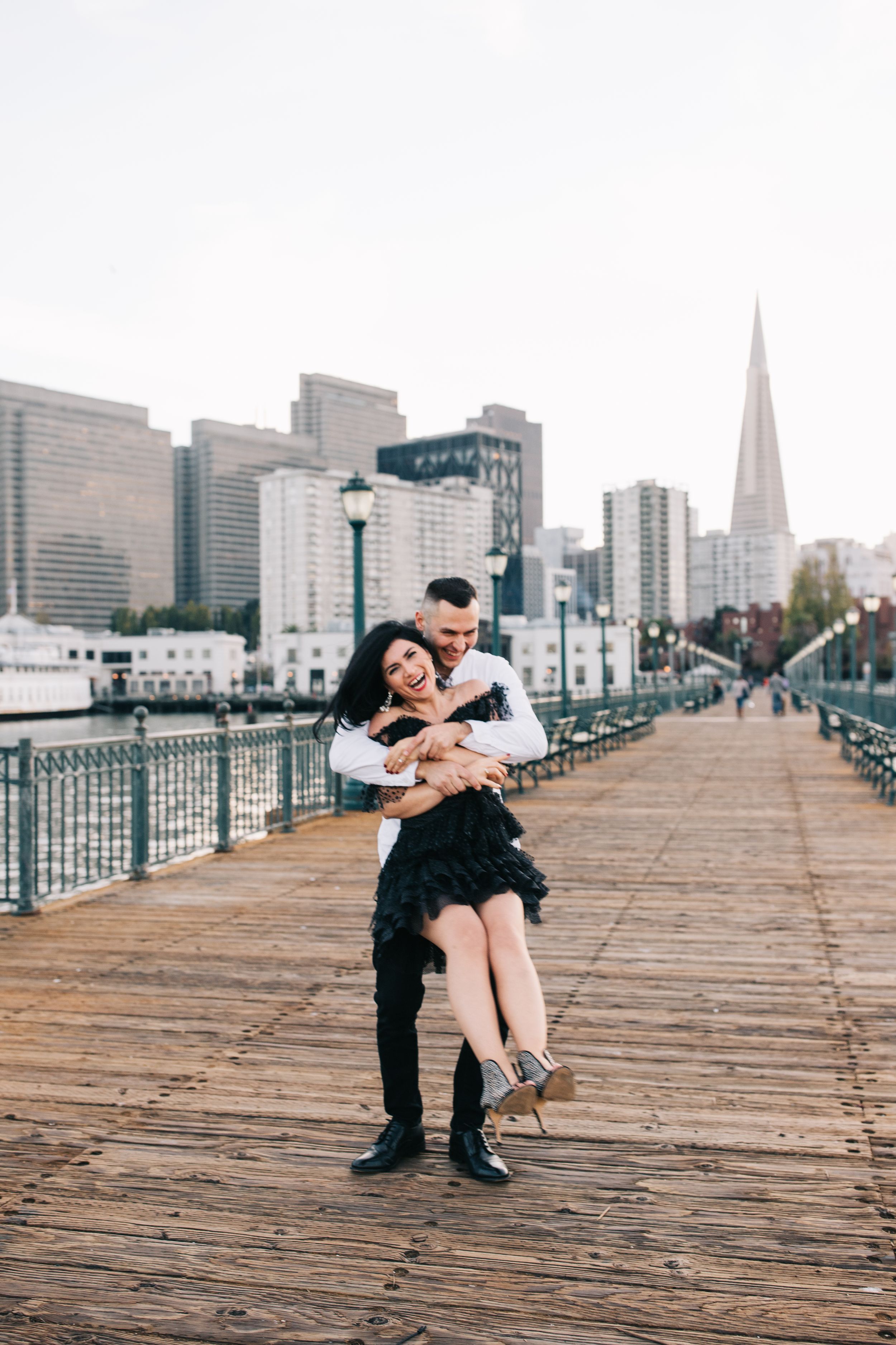 A romantic couple embraces on a wooden pier with the San Francisco skyline and Transamerica Pyramid in the background.