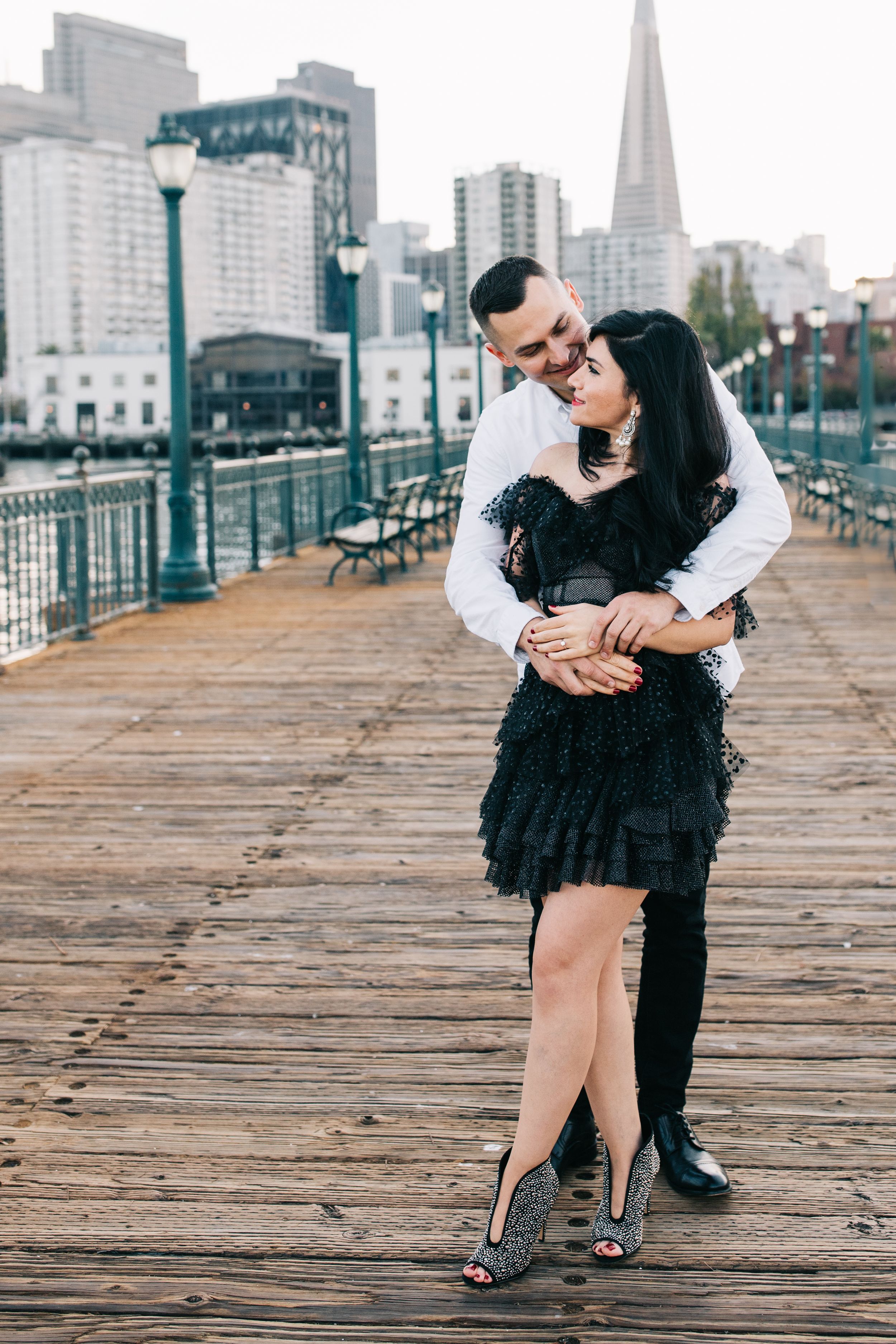 A romantic embrace on a wooden pier with the San Francisco skyline visible in the background.