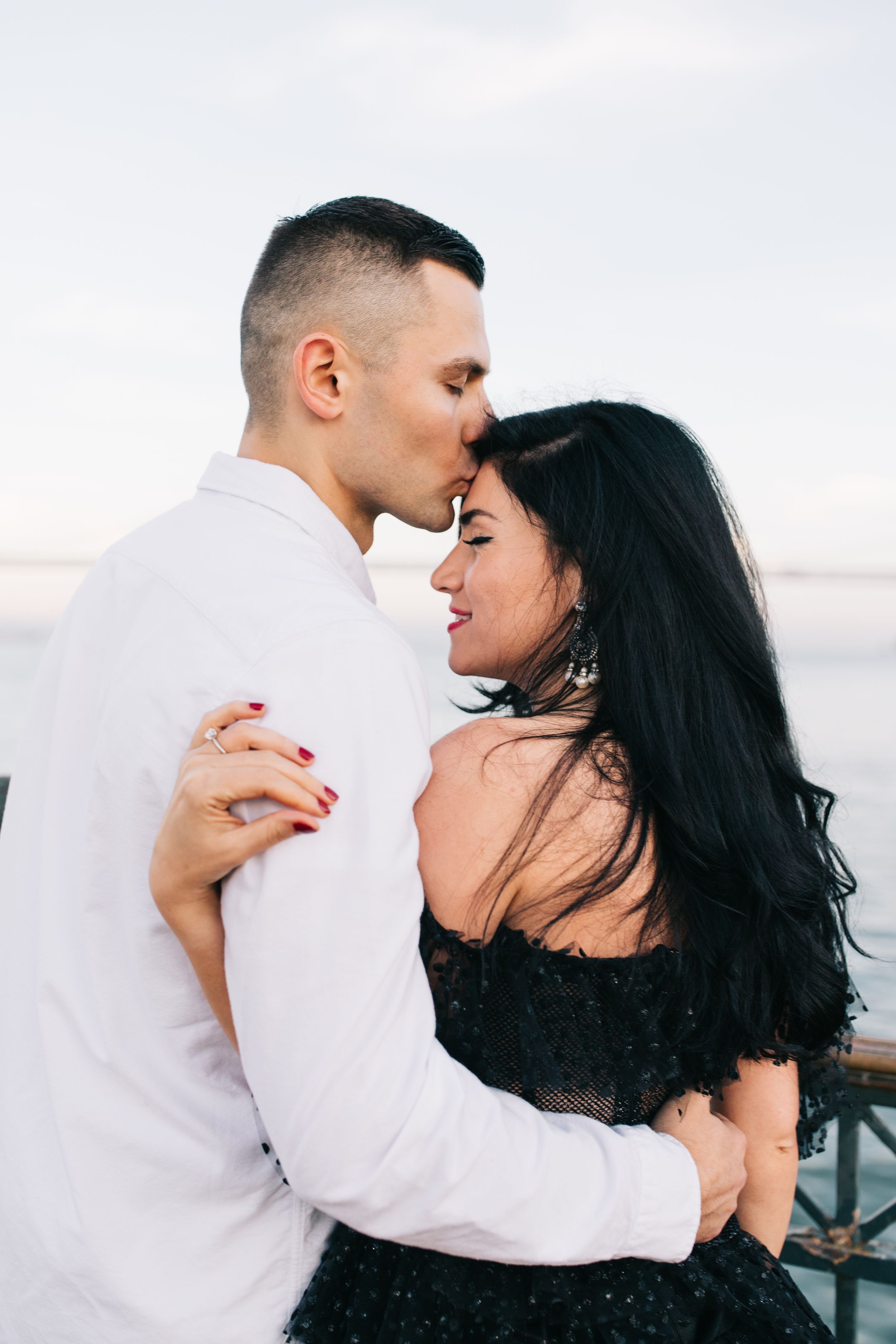 A couple shares an intimate moment on a waterfront with soft lighting and urban backdrop.