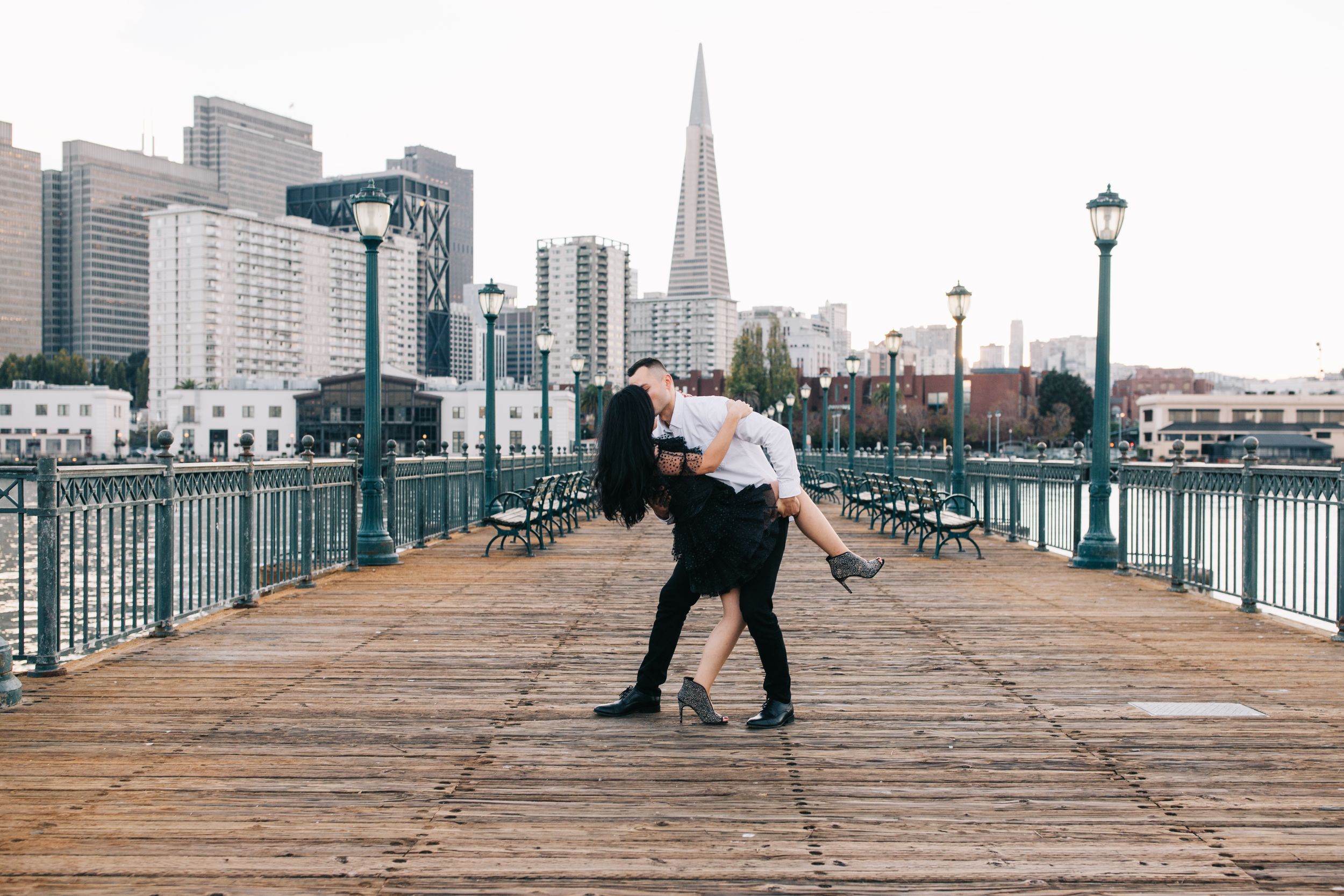 A couple dances romantically on a pier with the San Francisco skyline visible in the distance.