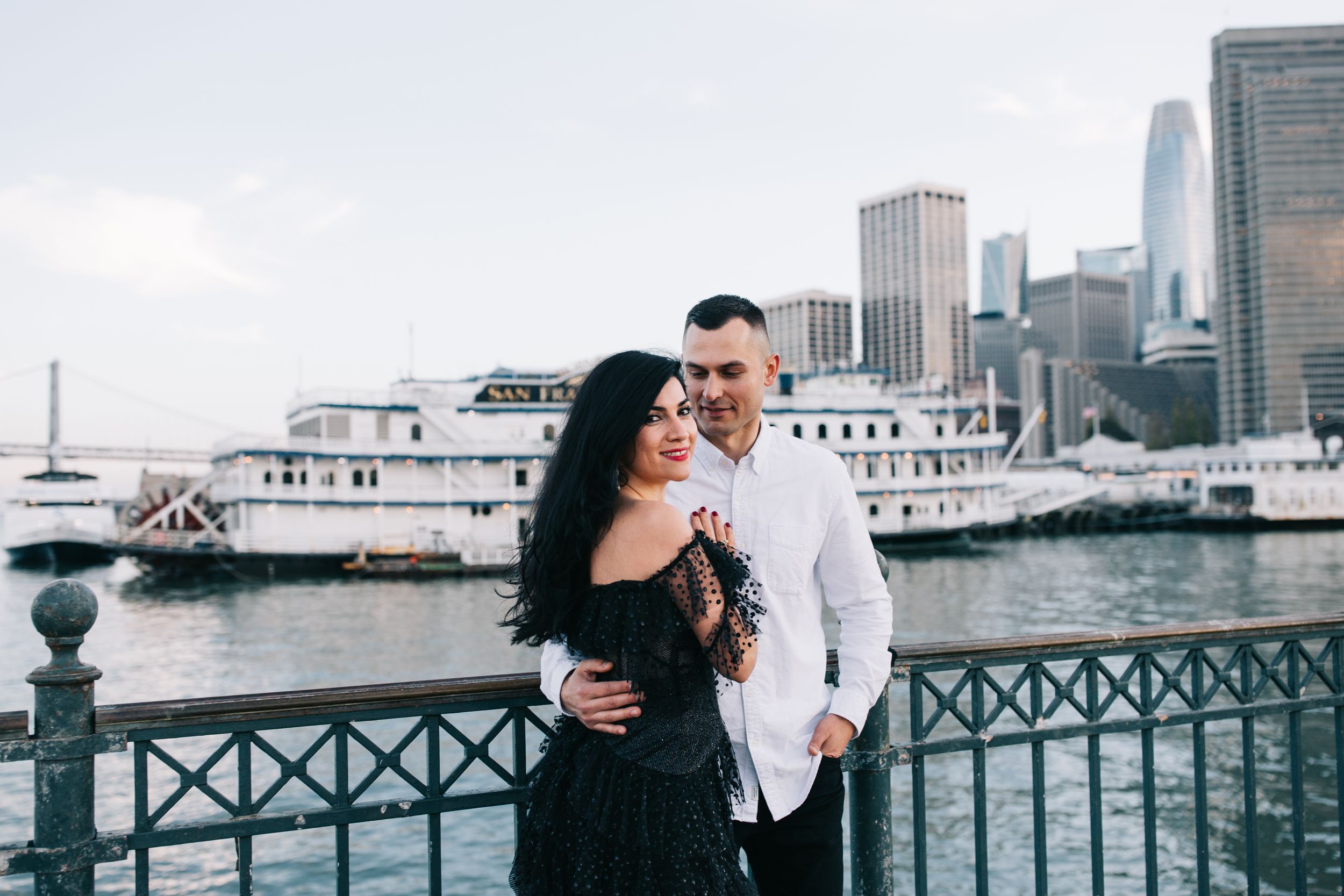 A couple poses along the San Francisco waterfront with boats and skyscrapers in the background.
